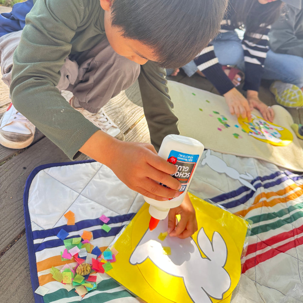 Child applying glue to a craft project with other people in the background.